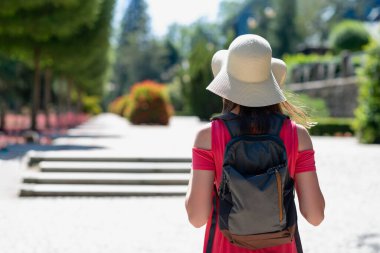 Geniş kenarlı şapkalı ve sırt çantalı bir kadının şehir parkında gezmesi, seyahat ve macera konsepti, yaz kentsel yaşam tarzı portresi