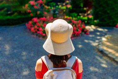 Geniş kenarlı şapkalı ve sırt çantalı bir kadın elinde çiçeklerle yaz bahçesinde dikiliyor, açık hava manzarasının tadını çıkarıyor, güneşli bir gün ve seyahat zamanı.