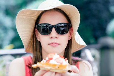 Geniş kenarlı şapkalı kadın elinde çilek ve kremayla taze waffle tutuyor, açık havada yaz tatlısı, kafede ya da fırında yemek yiyor, tatlı bir ikram sunuyor.