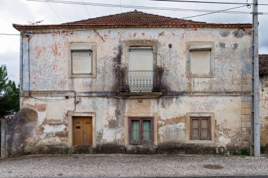 Terk edilmiş, balkonlu iki katlı köy evi yıpranmış cephe boyası, kiremitli çatı ve Portekiz 'de bulutlu gökyüzünün altındaki kaldırım taşı cadde boyunca kepenkli pencereler.