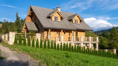 Taş aksanlı ahşap dağ kulübesi ve mavi gökyüzünün altındaki yeşil yamaçta geniş bir teras. Tepeleri ve ormanı olan kırsal alan huzurlu bir kırsal bölge yaratıyor, seyahat için ideal.