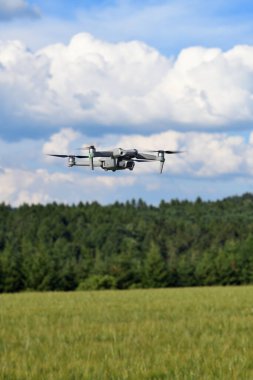 Uçan dron kuadkopteri orman ve tarla ile ön cephe görüntüsünü kapatıyor. Dijital kamerayla uçan İHA helikopter. Mavi gökyüzünde bir fotoğraf robotunun görüntüsü.