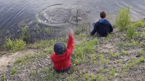 Kleiner Junge wirft Steine auf das schlammige Ufer eines Flusses — Stockvideo