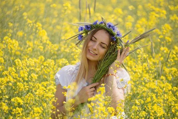 Menina com uma coroa de flores coloridas em sua cabeça, em um campo amarelo florido — Fotografia de Stock