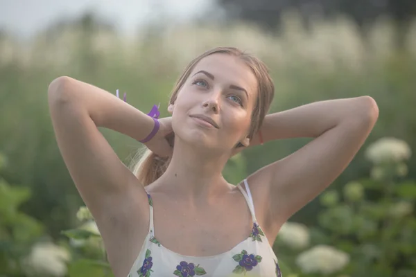 Jovem mulher bonita nos raios quentes do sol da noite andando em um campo verde com flores brancas . — Fotografia de Stock