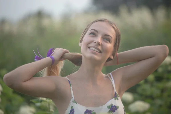 Young beautiful woman in the warm rays of the evening sun walking on a green field with white flowers. — Stock Photo, Image