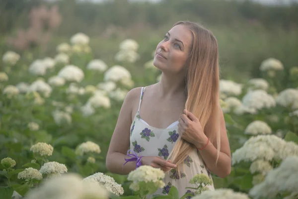 Νέοι όμορφη γυναίκα στις ζεστές ακτίνες του ήλιου που φιλτράρονται περπάτημα σε πράσινο φόντο με λευκά λουλούδια. — Φωτογραφία Αρχείου