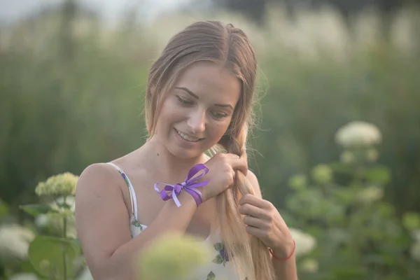 Jeune belle femme dans les rayons chauds du soleil du soir marchant sur un champ vert avec des fleurs blanches . — Photo