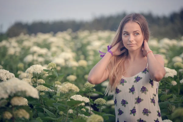 Joven hermosa mujer en los cálidos rayos del sol de la tarde caminando en un campo verde con flores blancas . — Foto de Stock