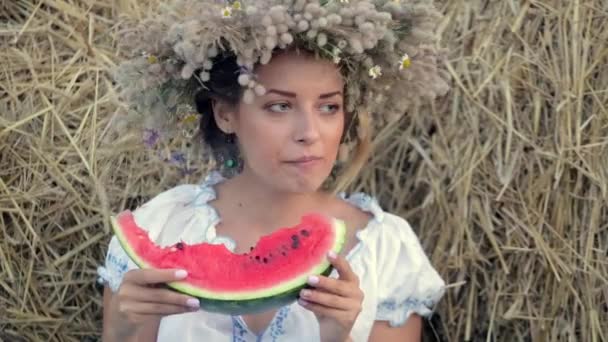 Молодая девушка в венке ест спелый арбуз возле стопок соломы — стоковое видео