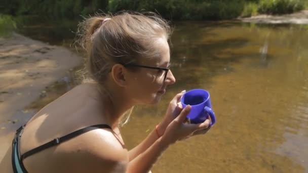 Flickan sitter vid vattnet med en kopp — Stockvideo