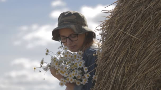 Girl in a hat with a bouquet of flowers is at stack of straw — Stock Video