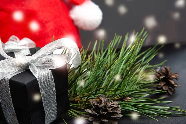 Black gift boxes with silver ribbons next to santa hat, green pine branch and fir cones with garland highlights. Closeup. Copy space