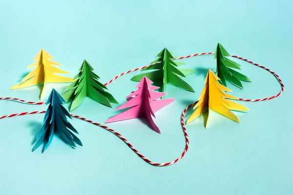 Multicolored paper trees stand against a blue background, and between them, like a path in the forest, a red and white Christmas thread is laid. Copy space. Top view. Mindfulness. Close up
