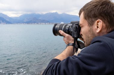 Fotoğrafçı, deniz ve dağların fotoğraflarını çekiyor. Yakışıklı bir yetişkin adam seyahat sırasında büyük kamerasını kullanır. Adam büyük bir kamera tutuyor ve ekrandan bakıyor..