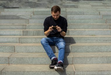 Elinde akıllı telefonu olan yetişkin bir adam taş merdivenlerde oturuyor ve mesajı yazıyor. Kot pantolon ve sweatshirt giyen bir adam cep telefonunu açık havada kullanıyor. Halk ve sosyal ağ kavramı.
