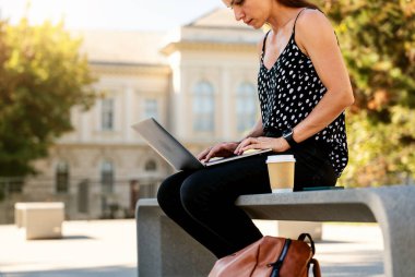 Şehir caddesinde bankta otururken dizüstü bilgisayar kullanan bir kadın. Fotokopi alanı olan kahverengi kağıt kahve fincanı..