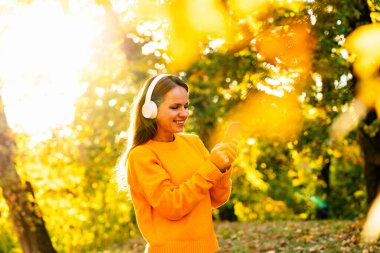 Sonbahar parkında yürürken akıllı telefon kullanan kablosuz kulaklıklı genç bir kadın.