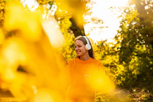 Güneşli sonbahar parkında kablosuz kulaklıklı akıllı telefon kullanan neşeli bir kadın.