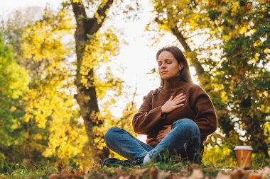 Ağaçlık alanda bağdaş kurmuş bir kadın oturuyor ve nefes egzersizi yapıyor. Stres atma ve akıl sağlığı.