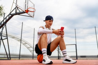 Basketbolun üzerinde oturan bayan sporcu sokak sahasında akıllı telefon kullanıyor.