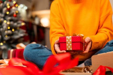 Sarı kazak giyen bir kadın elinde kırmızı renkli bir Noel hediyesi kutusu tutuyor..