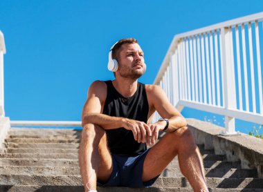 Spor yaptıktan sonra merdivenlerde dinlenen, kulaklık takan sağlıklı bir erkek portresi..