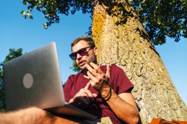 Çevrimiçi toplantı yaparken ağaçların yanında oturan alçak görüşlü şehirli adam..