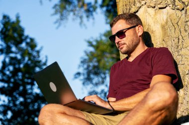 Yetişkin bir adam, ağaçların yanında otururken parkta dizüstü bilgisayarla çalışıyor..
