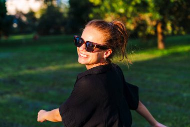 Şehir parkında yürürken omzunun üzerinden kameraya bakan neşeli bir kadın..