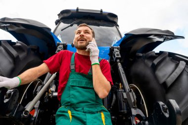 Açık hava traktörünün yanında telefonla konuşan düşük açılı tarım teknisyeni..