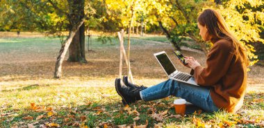 Sonbaharda sonbahar parkında dizüstü bilgisayar ve akıllı telefon kullanan bir kadın.