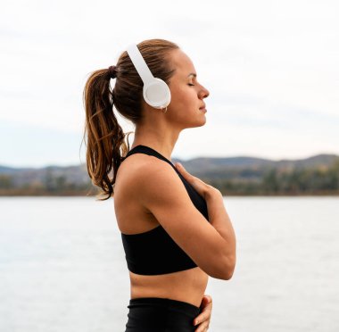 Kulaklıklı dikkatli bir kadın göğsünün üstünde el ele tutuşuyor. Nehir kenarında nefes alma egzersizi yapıyor..