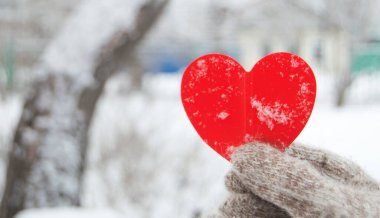 Bir kadının eli sıcak, örülmüş bir eldivende kırmızı bir kalp taşır. Şehir parkının bulanık arka planında, kar, kış, sevgililer günü konsepti, yakın plan.