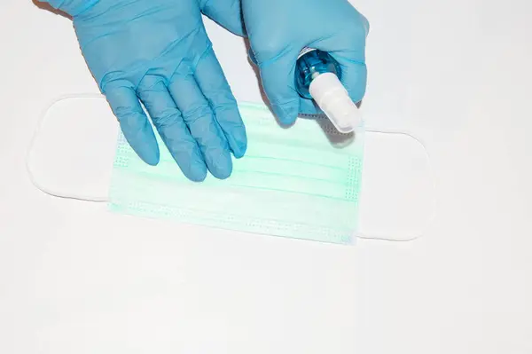 Mavi lateks eldivenli bir sıhhiyenin ellerinde beyaz bir arka planda cerrahi maske ve antiseptik sprey bulunur. Korunma kavramı