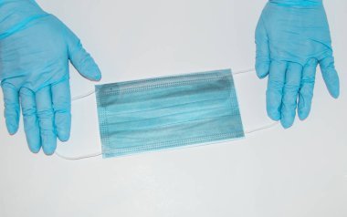 Mavi lateks eldivenli bir sıhhiyenin elleri beyaz arka planda kulak ilmiği olan bir cerrahi maske tutar. Korunma kavramı