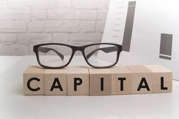 The word CAPITAL is written on wooden cubes standing on the white surface of the table, the GLASSES are on the cubes — Stockfoto