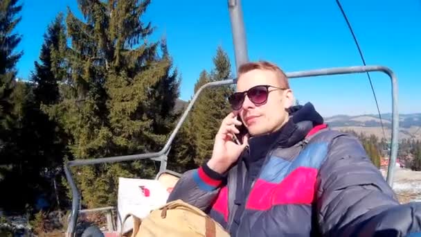 Hombre guapo monta un teleférico contra el fondo de un bosque de invierno y habla por teléfono. — Vídeo de stock