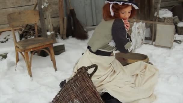 Egy vörös hajú lány etnikai ruhában egy régi falusi ház hátterében áll egy téli napon.. — Stock videók