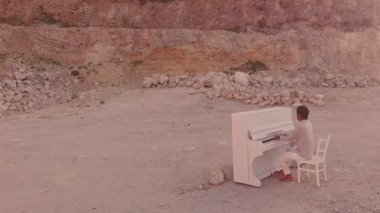 Genç adam sarı kumdaki beyaz bir piyanoda sarı taşlardan oluşan bir arka planda çalıyor. Hava görünümü
