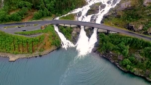 Uma bela cachoeira poderosa flui de uma montanha alta sob a ponte e flui para um grande rio. Vista aérea. Noruega. — Vídeo de Stock