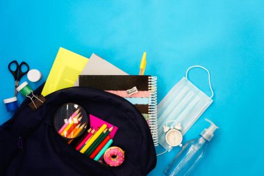 COVID-19 'un okul önlenmesine, okul malzemeleriyle dolu sırt çantasına, mavi kumaş maskesine ve mavi arka planda dezenfektan jeline geri dönüyoruz..