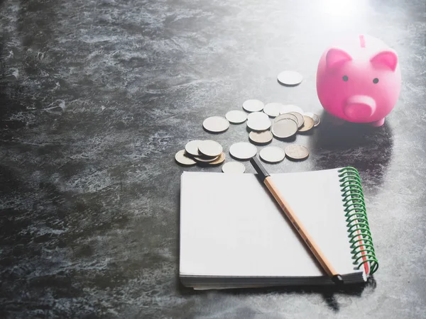 A piggy bank stands on a table with scattered coins and a notepad