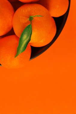 Turuncu arka planda mandalinalar ve kâsenin üzerinde yapraklarla mandalina.