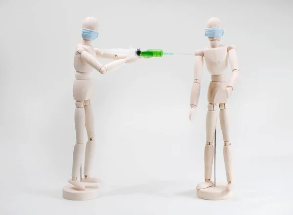 The concept of vaccination or injections for protection from viruses and bacteria. A wooden model of a masked man vaccinates a green vaccine to another wooden person. neutral background.shoulder injection