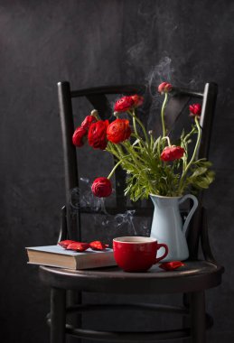 vintage still life on a black chair against a black background, red ronunculus in a blue jug, a red cup of tea with smoke, a blue book and candies in a red wrapper, dark photo