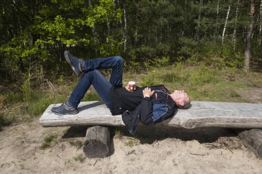Ormanın ortasında seyahat ettikten sonra bankta dinlenen orta yaşlı bir gezgin, güneşli bir gün, elinde bir kupa soğuk Starbucks kahvesi tutan yaşlı bir vatandaş, as, Belçika, 21 Mayıs 2021