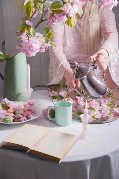 Young woman pours coffee into a cup on a background of sakura flowers and colored macaroon cakes, retro style life, spring season — Stock Photo, Image