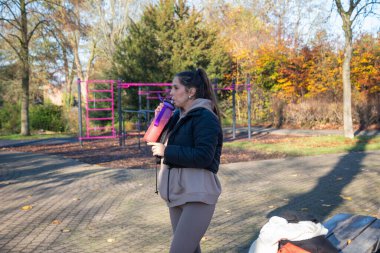Renkli bir şişeden su içen genç bir kadın, güneşli bir parktaki güneşli bir spor salonunda antreman yaptıktan sonra serinliyor ve susuz kalıyor.