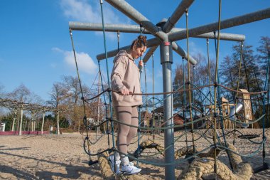 Atletik bir kadın, açık mavi gökyüzü altında denge ve fiziksel aktiviteye odaklanarak, kumlu bir oyun alanında zorlu bir halat köprü tırmanma yapısında geziniyor.